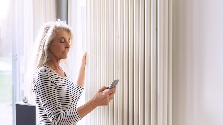 Strom- und Heizkosten senken Eine Person steht mit einem Smartphone in der Hand vor ihrer Heizung.